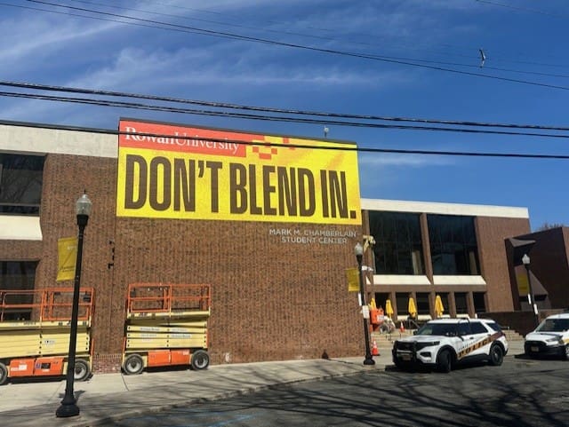 University building with "Don't Blend In" banner.